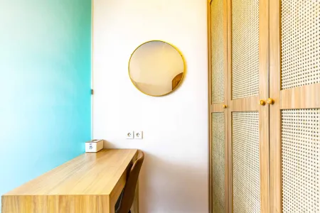 Modern hotel bedroom interior with wooden desk, rattan wardrobe, and gold round mirror against turquoise wall.